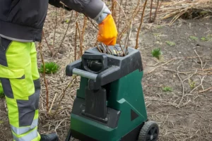 Henkilö syöttää oksia vihreään oksasilppuriin pihalla, maassa näkyy risuja ja kasvustoa.