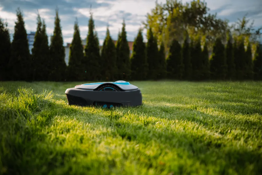 Robottiruohonleikkuri liikkuu vihreällä nurmikolla, taustalla korkeita puita ja sininen taivas.